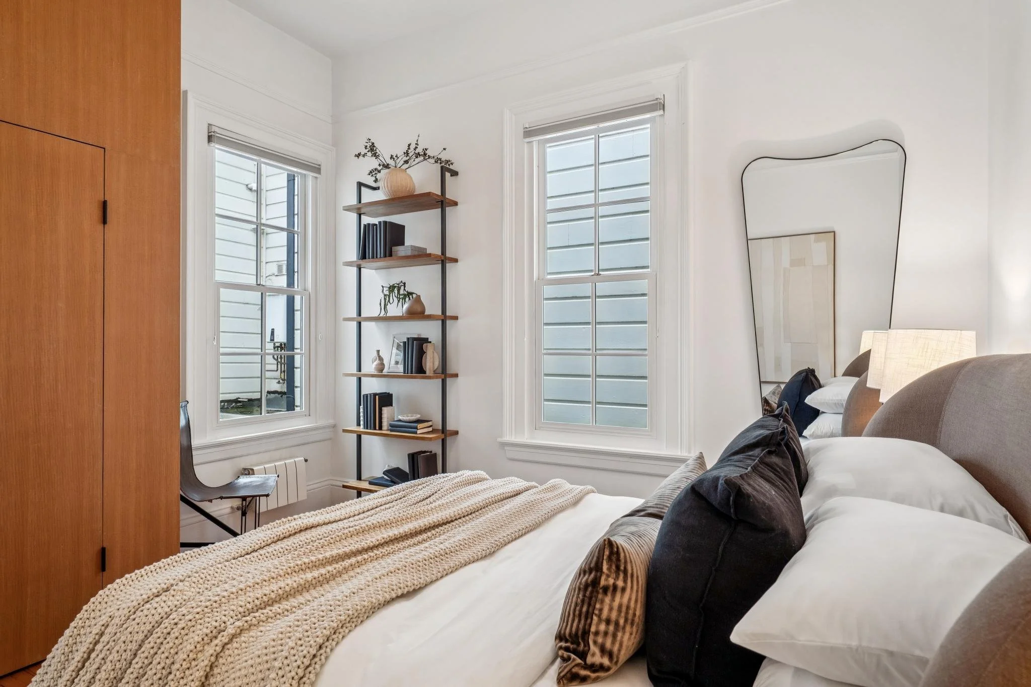 A cozy bedroom with a bed, throw pillows, a knitted blanket, two windows, a black chair, a wooden shelf with books and decor, and a large mirror on the wall.