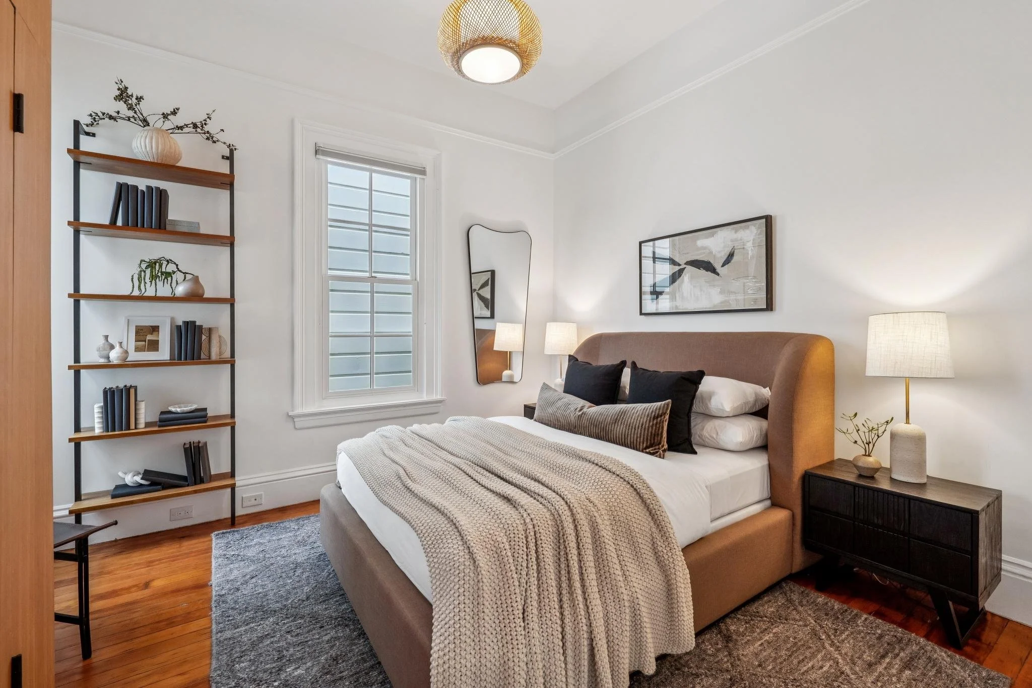 A cozy bedroom with a brown upholstered bed, black and beige pillows, and a cream knitted blanket. There is a wooden nightstand with a ceramic lamp and small plant, a rectangular mirror on the wall, a window with horizontal blinds, a wall art above t