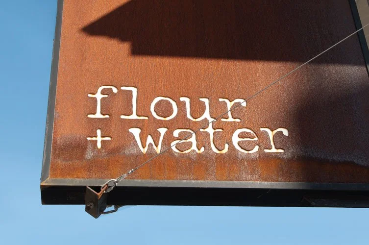 A rusty metal sign that reads 'flour + water' with a wire attached at the bottom left corner, set against a clear blue sky.
