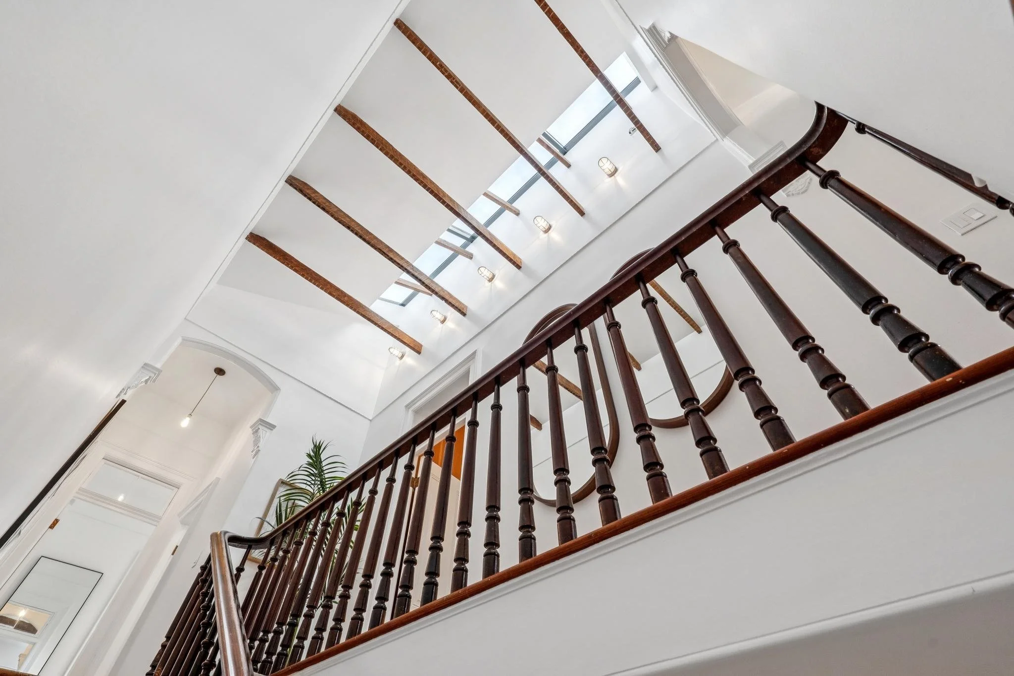 Interior view of a house staircase and ceiling, featuring wooden railing, white walls, ceiling lights, and exposed wooden beams on the ceiling.