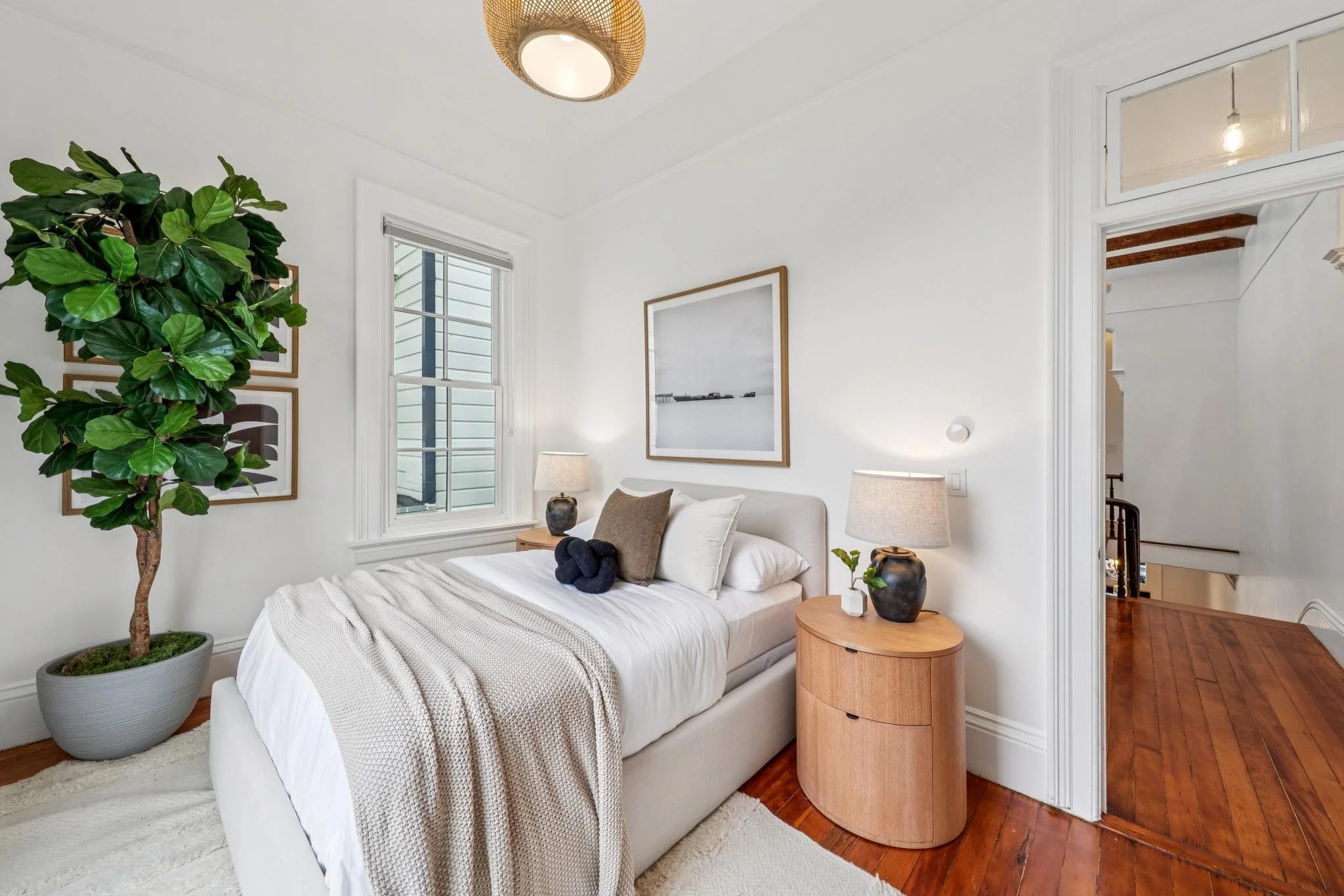 A cozy bedroom with white walls, a cream bed with pillows and a blanket, wooden side tables with black lamps, a large potted plant, a window, and framed art, with hardwood floors and an open doorway leading to a staircase.