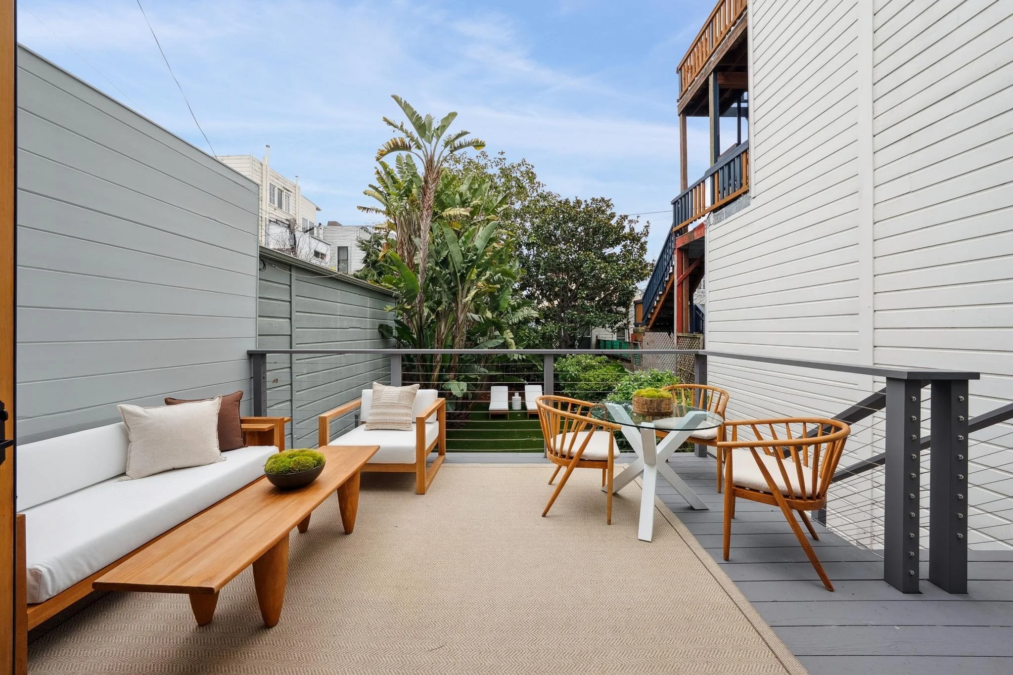 Outdoor patio with white cushioned sofa, two wooden armchairs, a glass-top table with wooden chairs, beige rug, potted plants, and green trees in background.