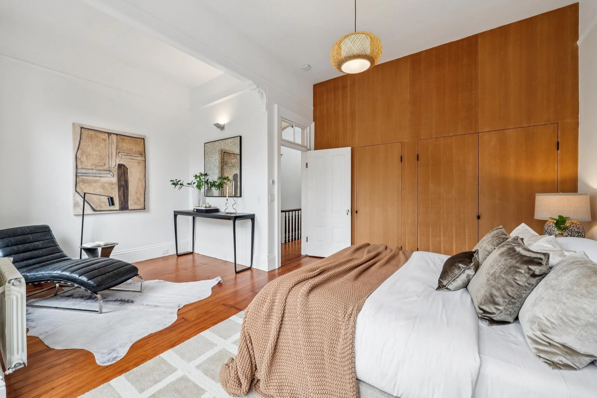 A modern bedroom with a large bed covered in white bedding and beige pillows, a brown throw blanket, a side table with a lamp, a black lounge chair, a white rug, wooden flooring, a wooden accent wall, a black console table with a mirror and plant, a 