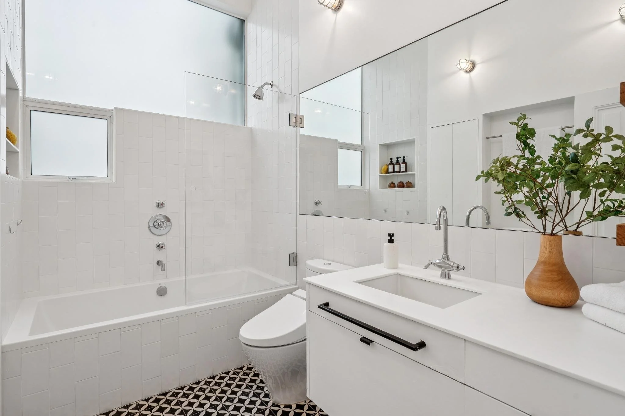 Modern white bathroom with a bathtub, toilet, sink, large mirror, frosted windows, and decorative plants.