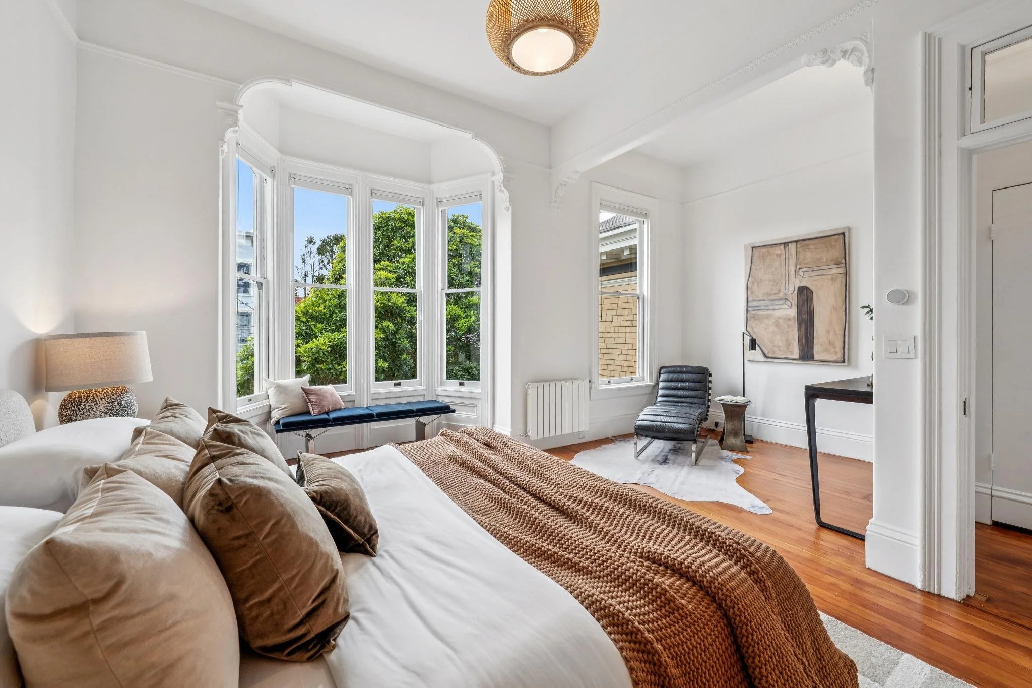 Bright bedroom with large bay window, white walls, hardwood floors, and modern furnishings including a bed with beige pillows and a brown throw, a black chair, a cowhide rug, and abstract artwork on the wall.