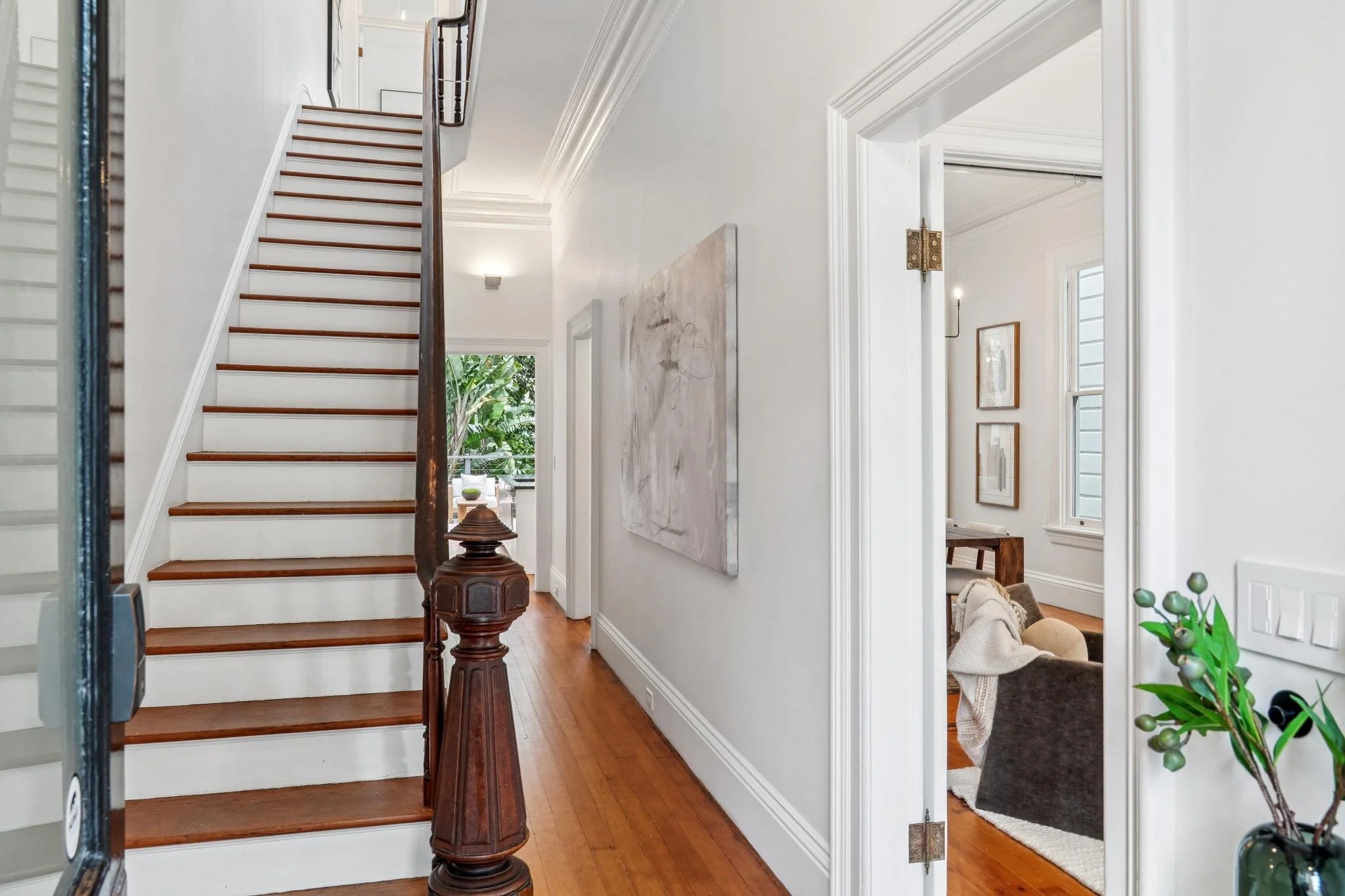 Interior view of a house showing a staircase with wooden steps and a dark wooden handrail, a hallway with white walls and baseboards, a large abstract painting on the wall, and a doorway leading to a living room with windows, framed art, a chair, and
