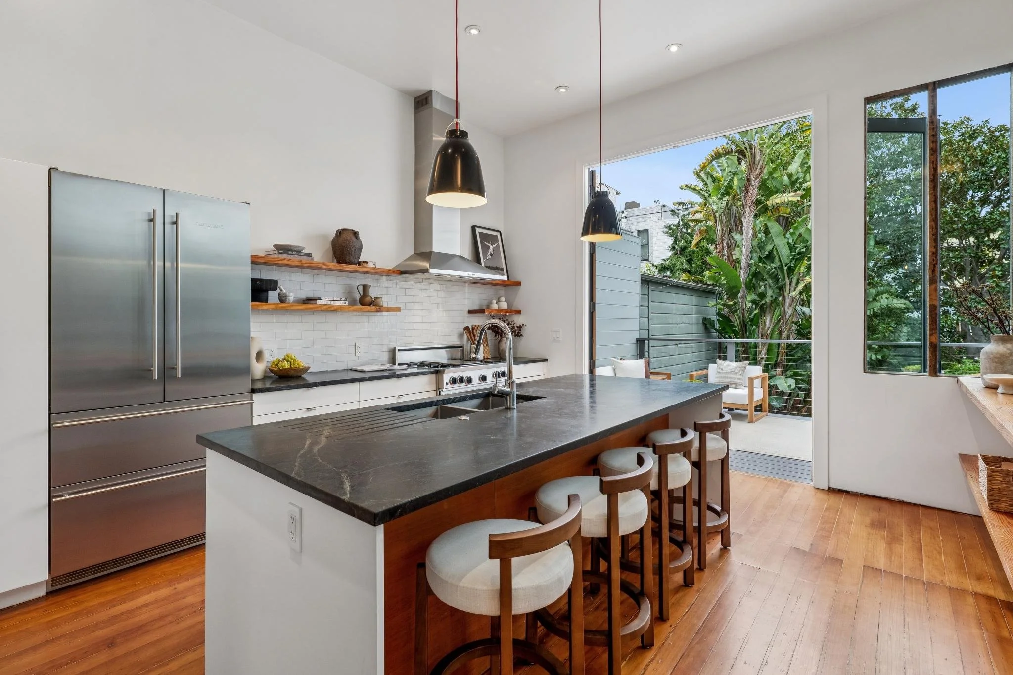 Modern kitchen with black marble island, wooden bar stools, stainless steel refrigerator, open shelves, white cabinetry, and sliding glass door leading to outdoor patio with greenery.