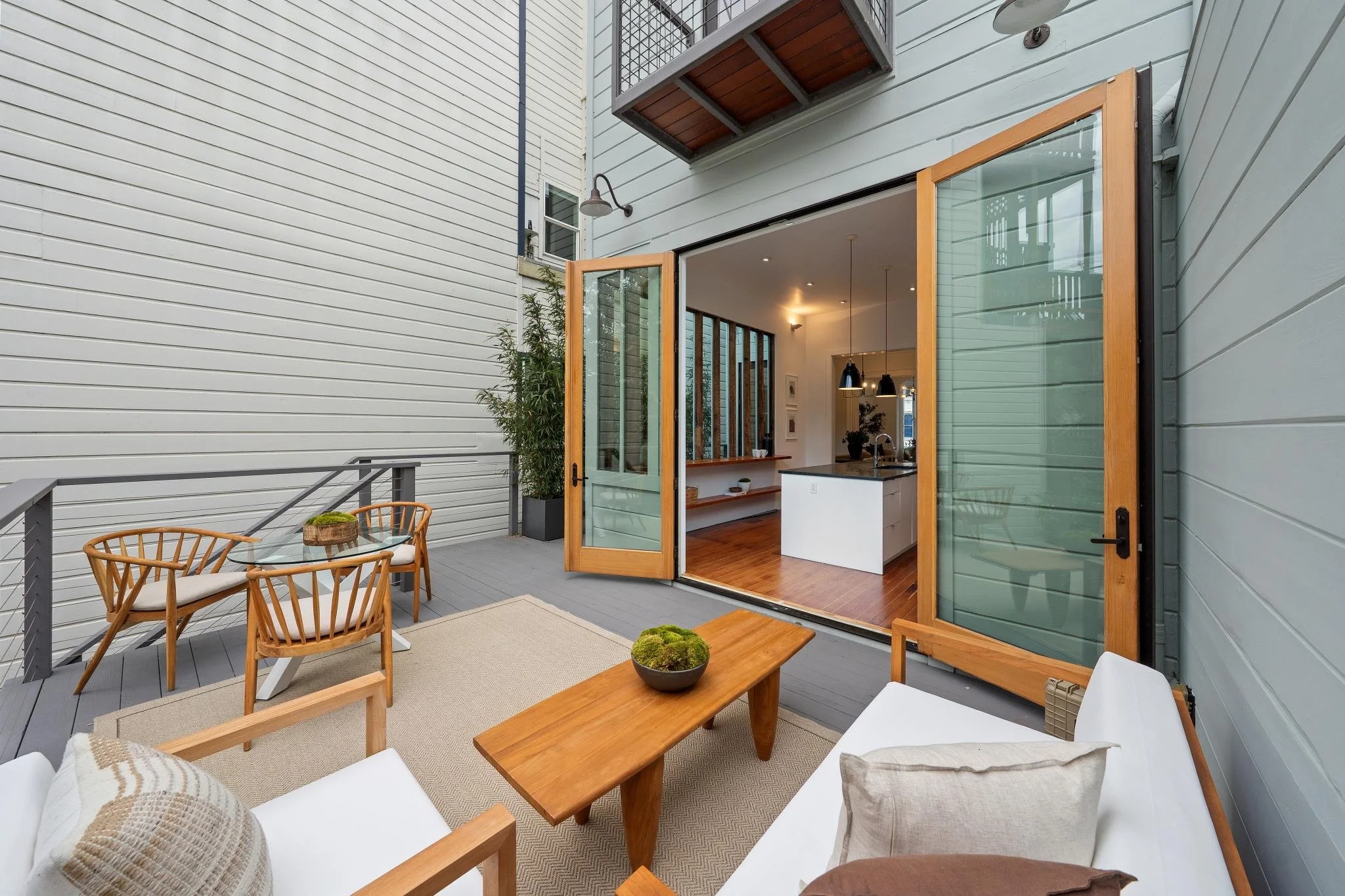 Outdoor deck area with wooden furniture, including a glass table with two chairs, a small wooden coffee table with a plant, and a white sofa with cushions. The deck opens to an indoor living space through wooden-framed glass doors.