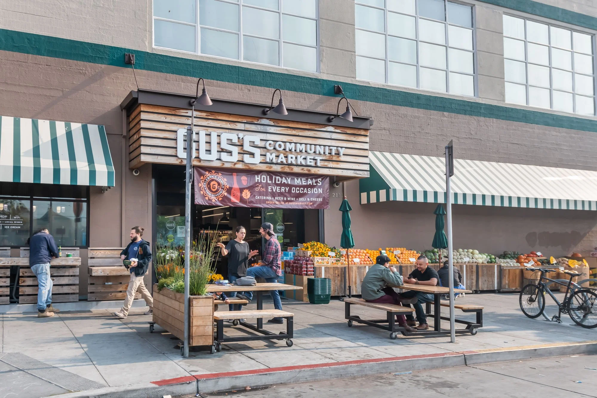 People shopping at and sitting outside a community market with a sign that reads 'GUSS Community Market,' featuring fresh produce and an outdoor seating area.