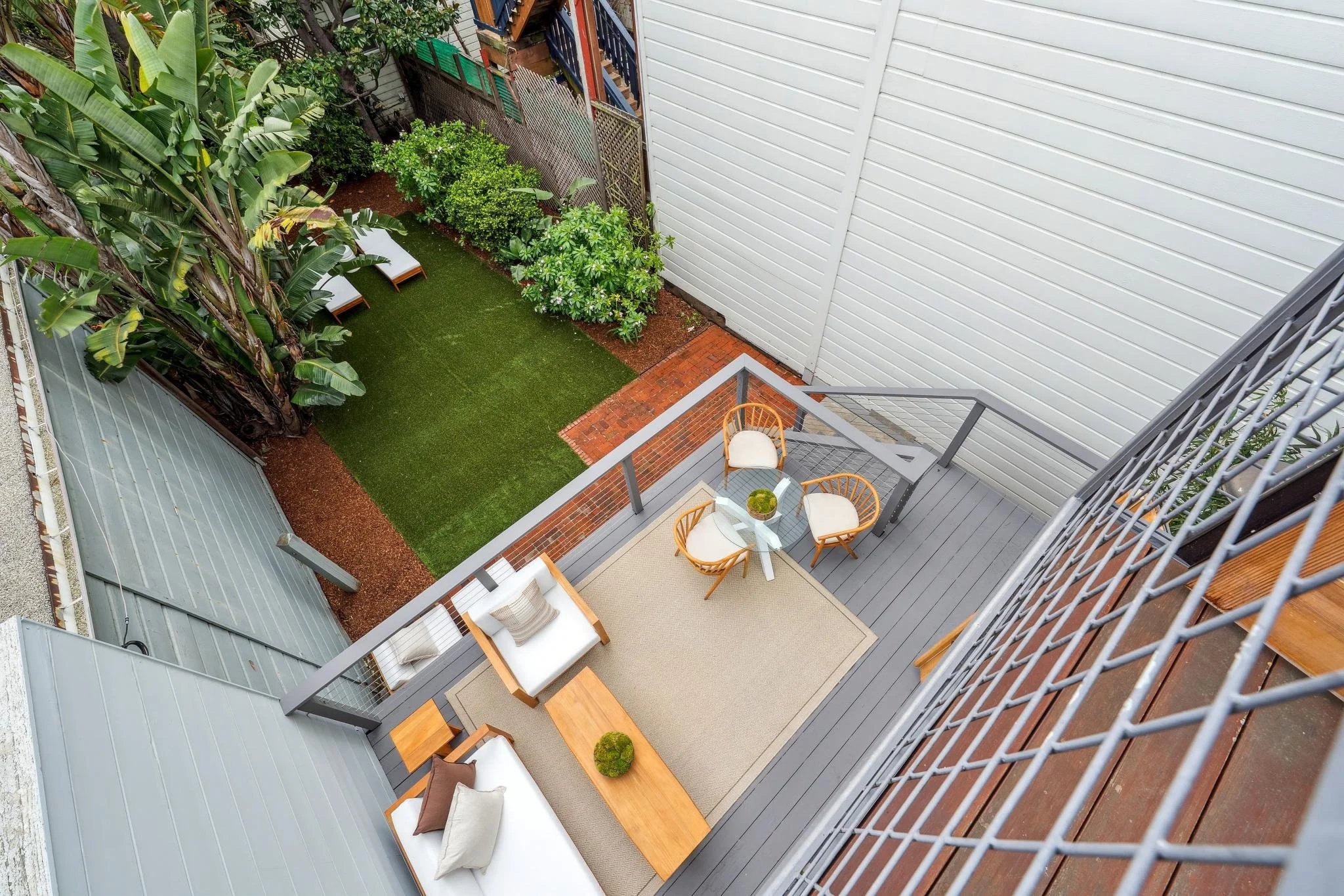 A small outdoor balcony with a seating area including a white cushioned chair, a sofa with pillows, a wooden coffee table, and a round glass table with three wooden chairs around it. The balcony has gray flooring and is enclosed by gray railings. Bel