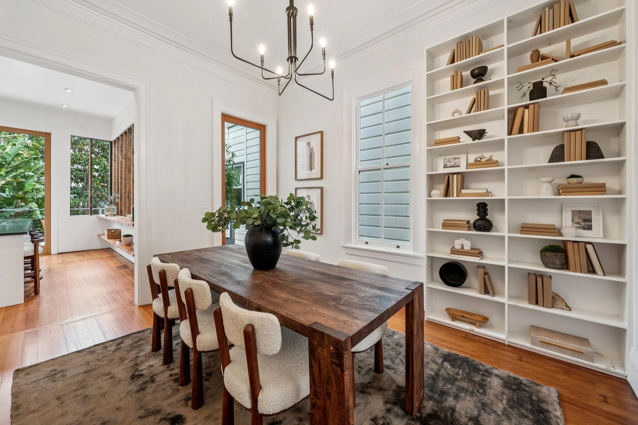 A dining room with a wooden table, six upholstered chairs, a black vase with greenery, large windows, a white bookshelf with decorative items, and light-colored hardwood flooring.