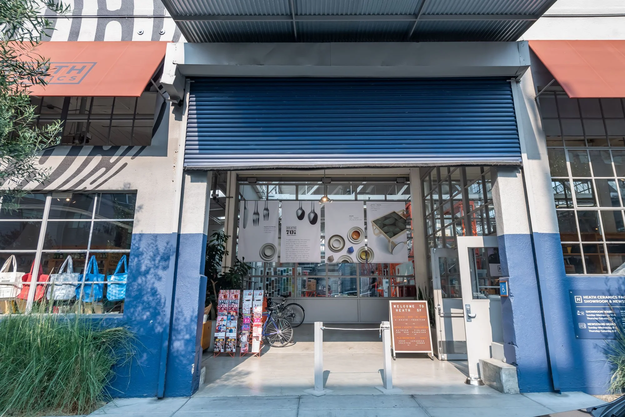The exterior of a showroom with a partially open blue metal shutter, featuring large windows displaying kitchenware posters, assorted bags on a rack, bicycles, and a welcome sign at the entrance.