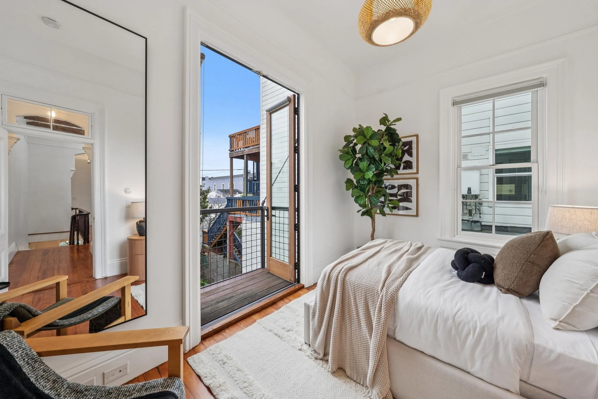 A cozy bedroom with white walls, hardwood floor, and a balcony door leading outside. There is a white bed with neutral pillows and a blanket, a tall plant, and wall art. A mirror reflects the room and hallway.