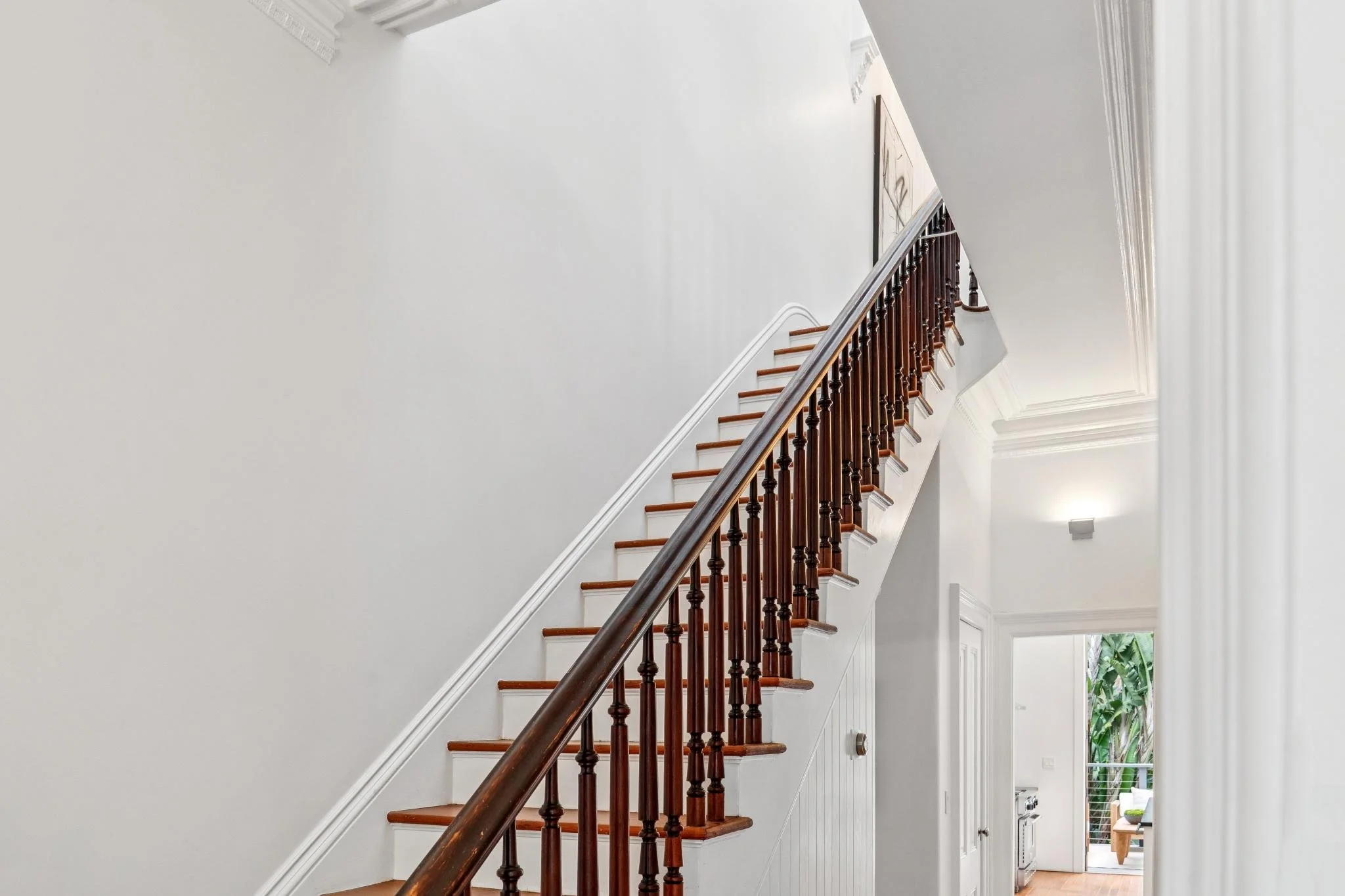 Interior view of a staircase with wooden handrail and spindles, white walls, and decorative crown molding. Natural light coming from a door leading to an outdoor balcony with balcony railing and greenery.
