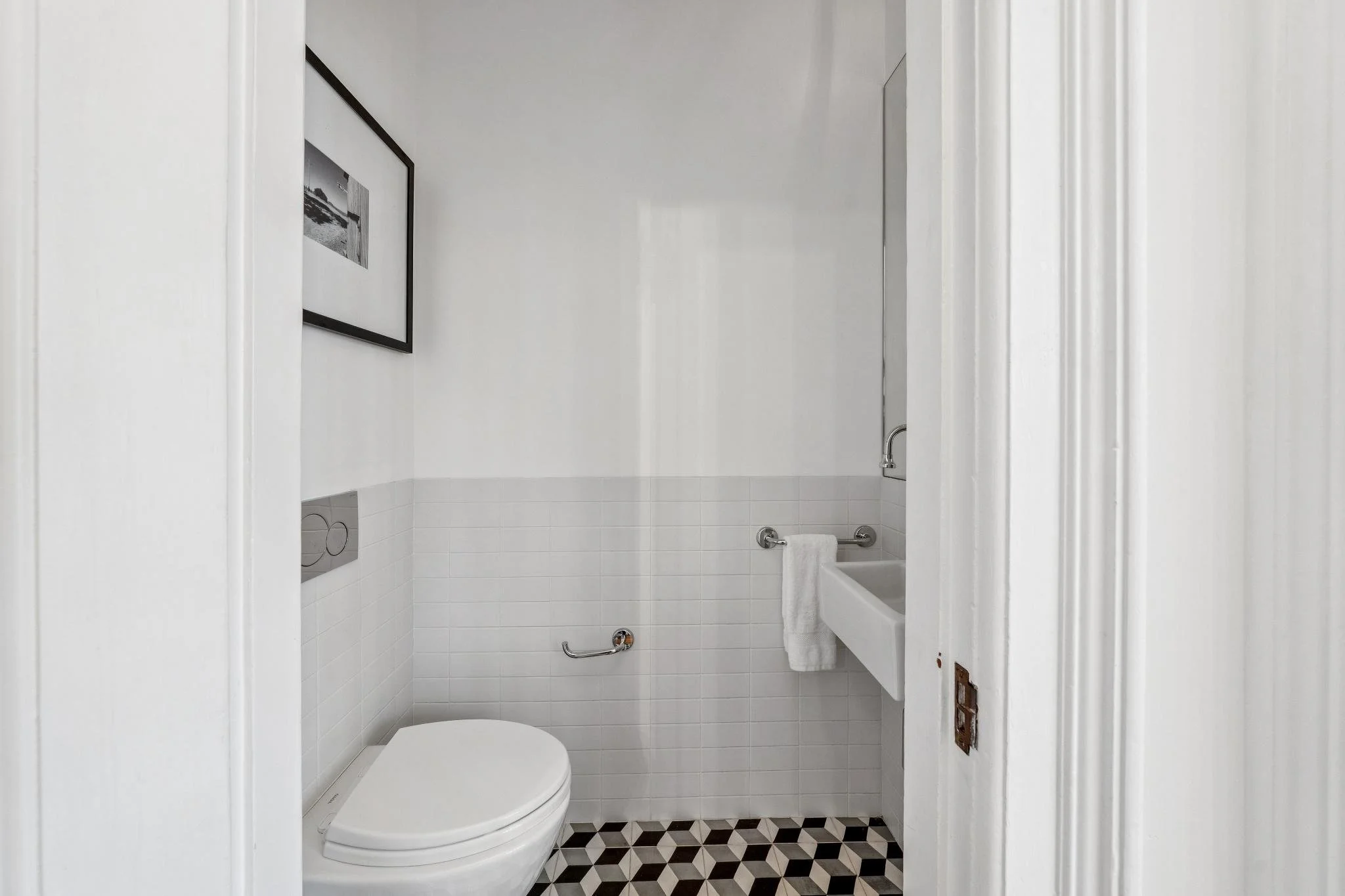 Small bathroom with white tile walls, a wall-mounted toilet, a small white sink with a towel, and a black-and-white framed picture on the wall.