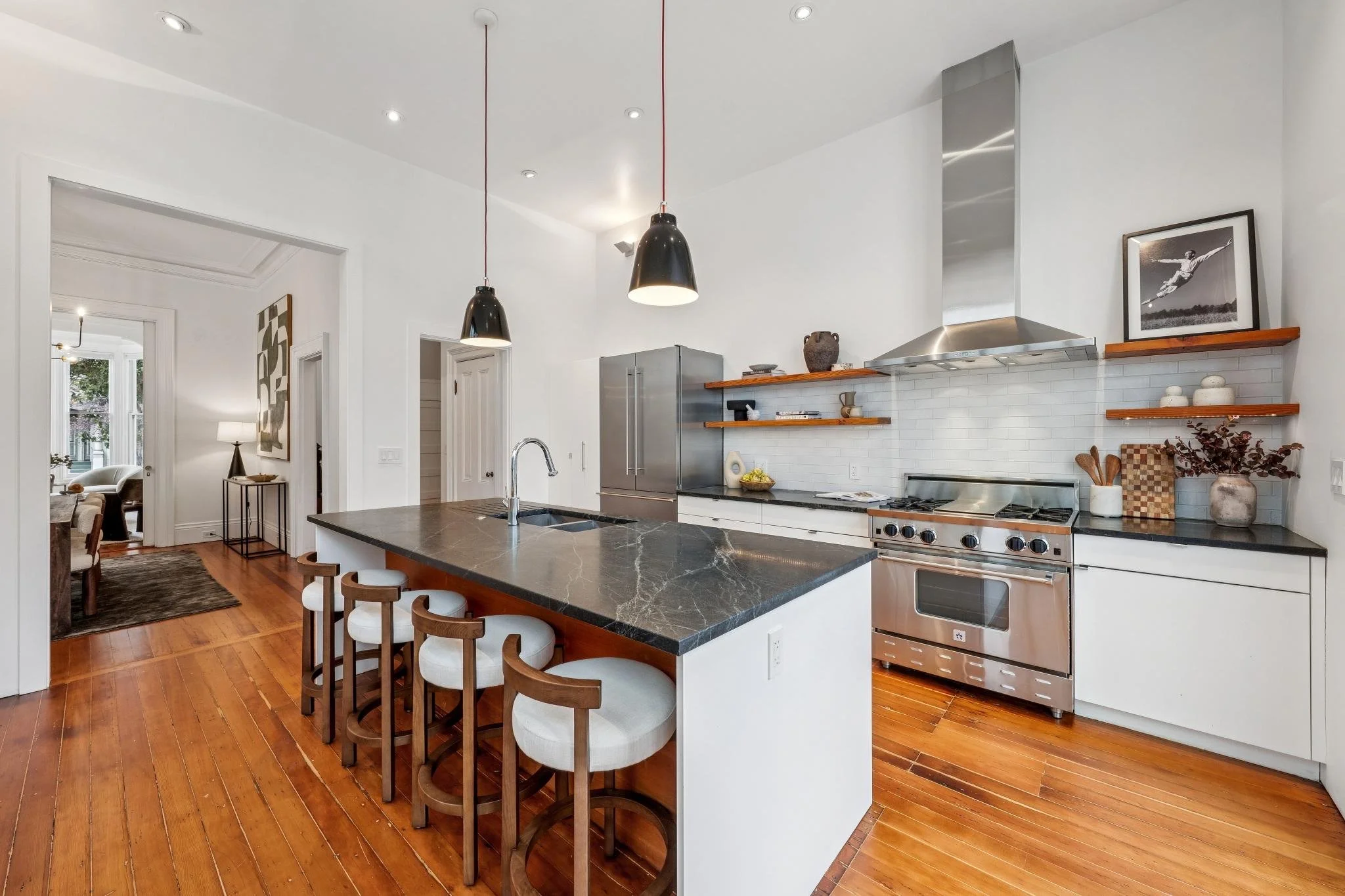 Modern kitchen with white walls, wooden floors, black pendant lights, stainless steel appliances, open wooden shelves, and a black marble kitchen island with four chairs.