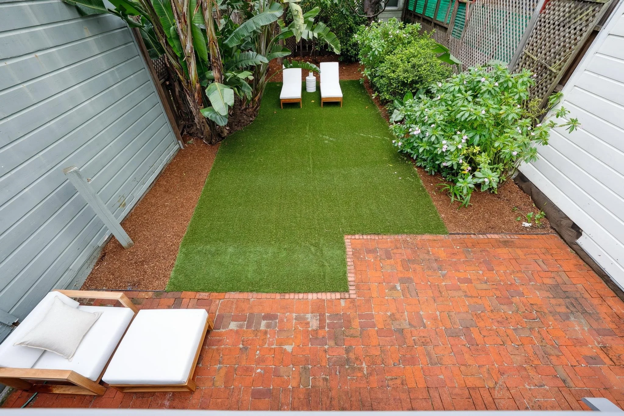 A backyard scene with brick patio, green artificial turf, two white outdoor chairs, and lush plants, including bananas and flowering bushes.