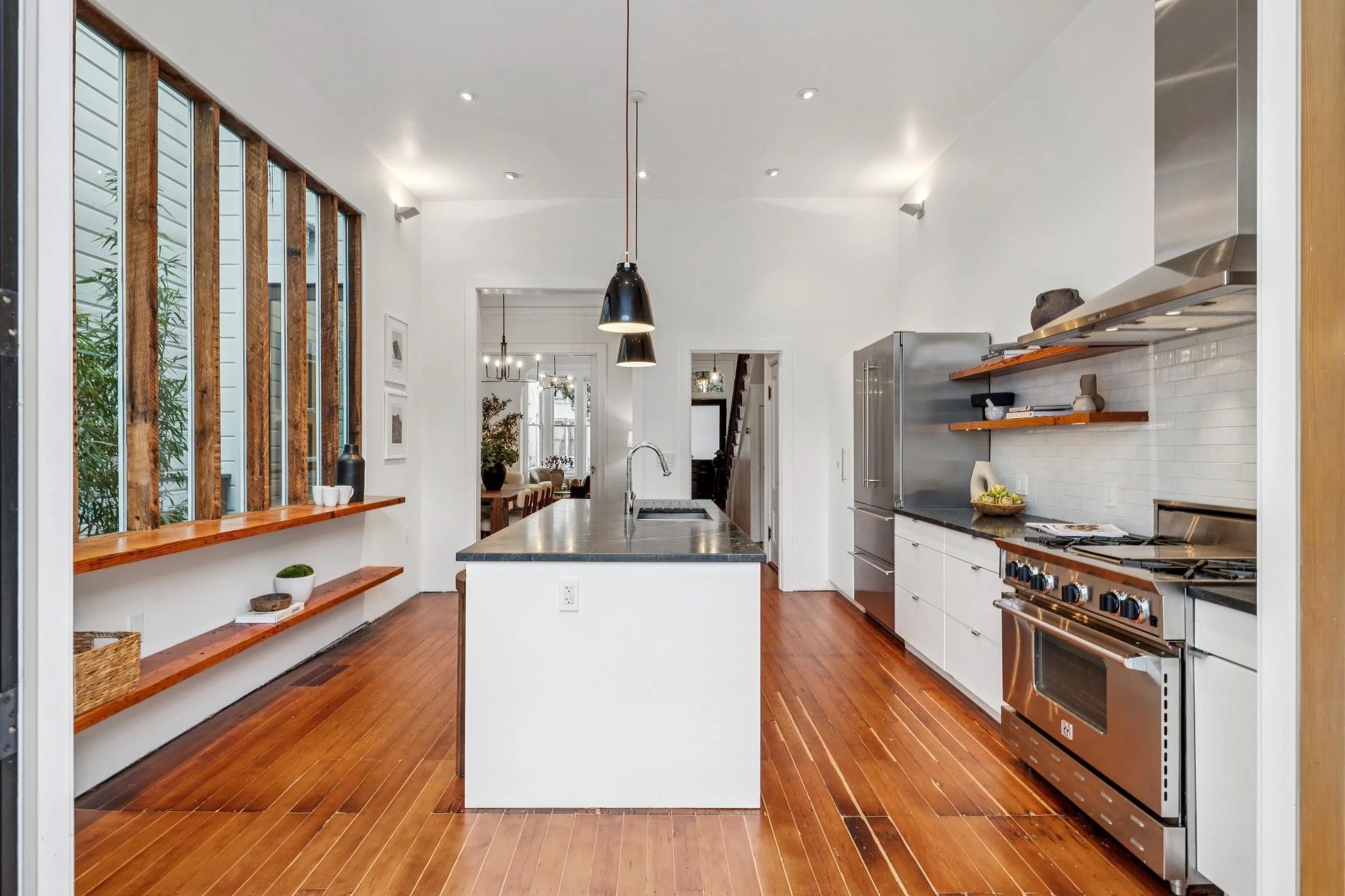 Modern kitchen with white cabinets, stainless steel appliances, black countertop island, wooden shelves, and hardwood floors. Large windows on the left letting in natural light.