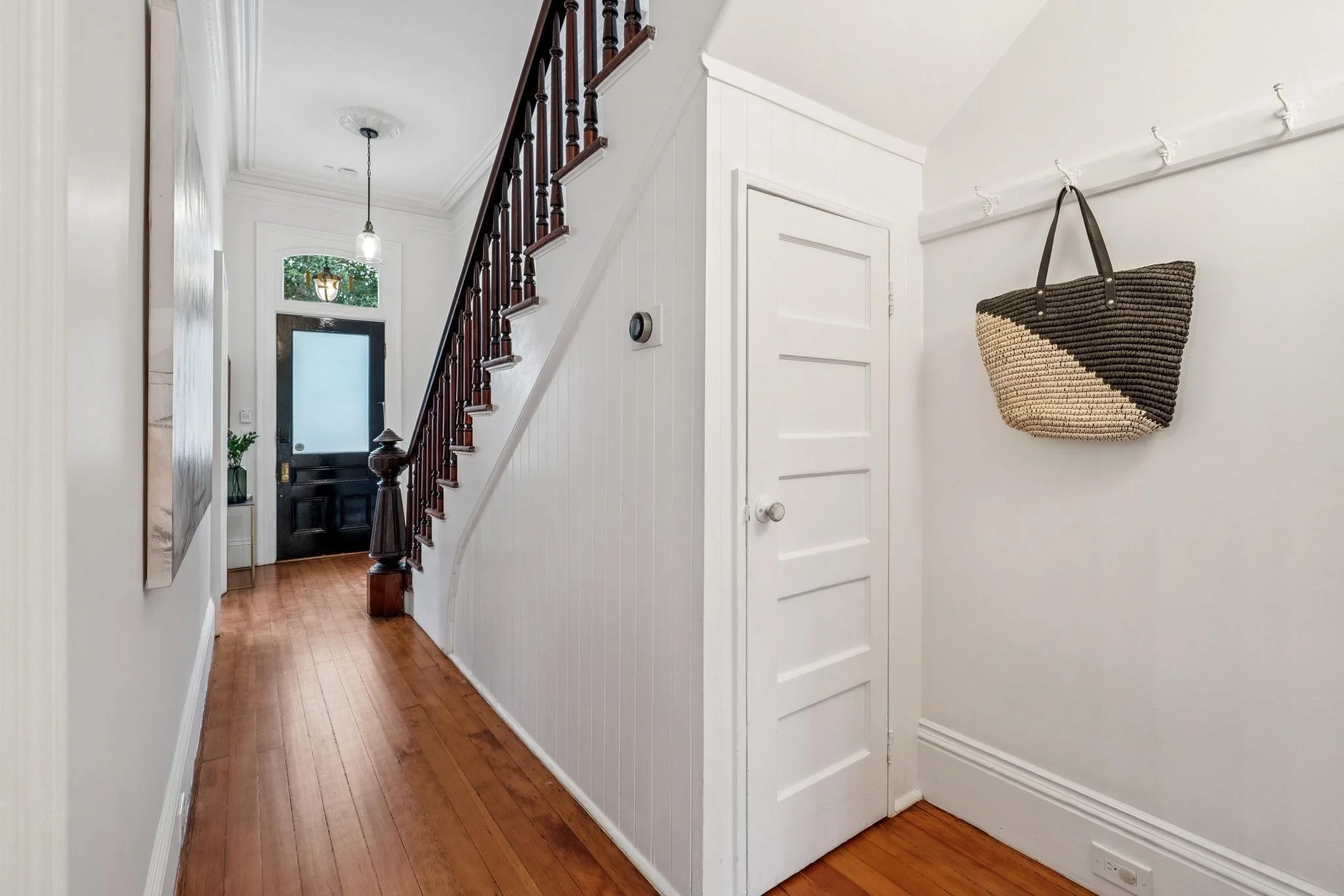 Entryway with wooden floor, white walls, staircase with dark wooden railing, front door with frosted glass window, wall-mounted basket with black strap, small side table with plant, hanging light fixture