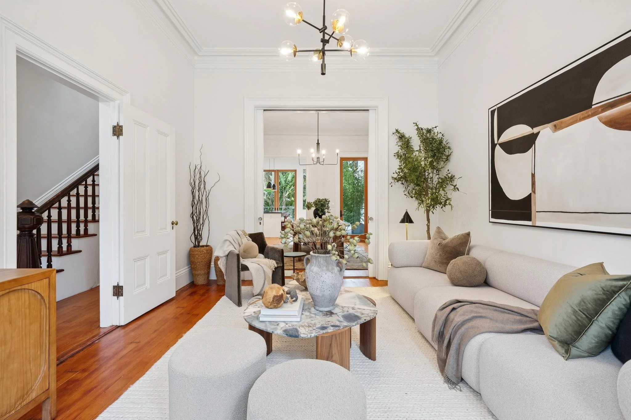 Living room with white walls, beige couch with pillows, abstract wall art, wooden floors, and a decorative marble coffee table with vases and books.