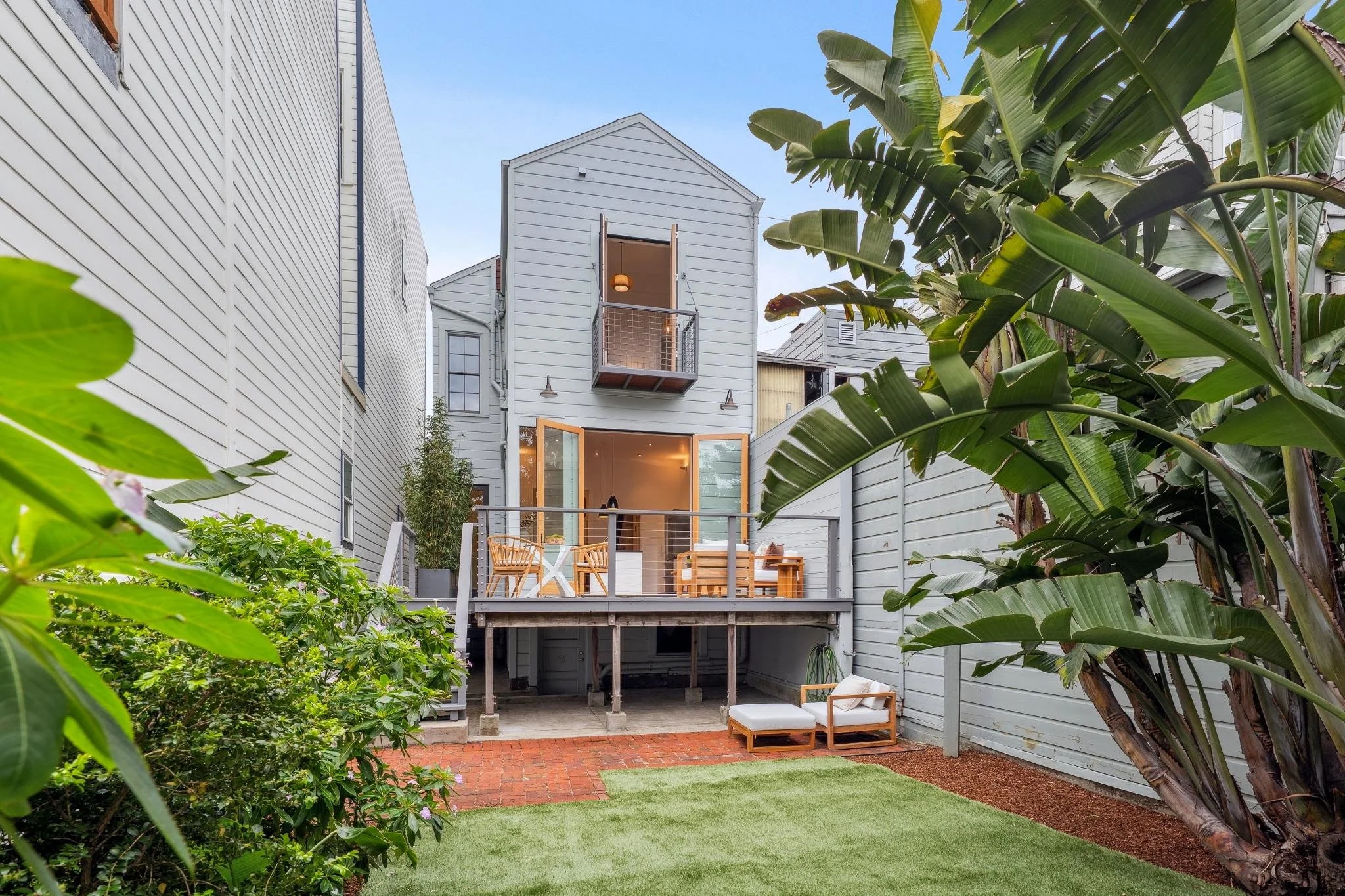 Backyard with a small lawn, a patio with outdoor furniture, and a gray house with large windows and a balcony.