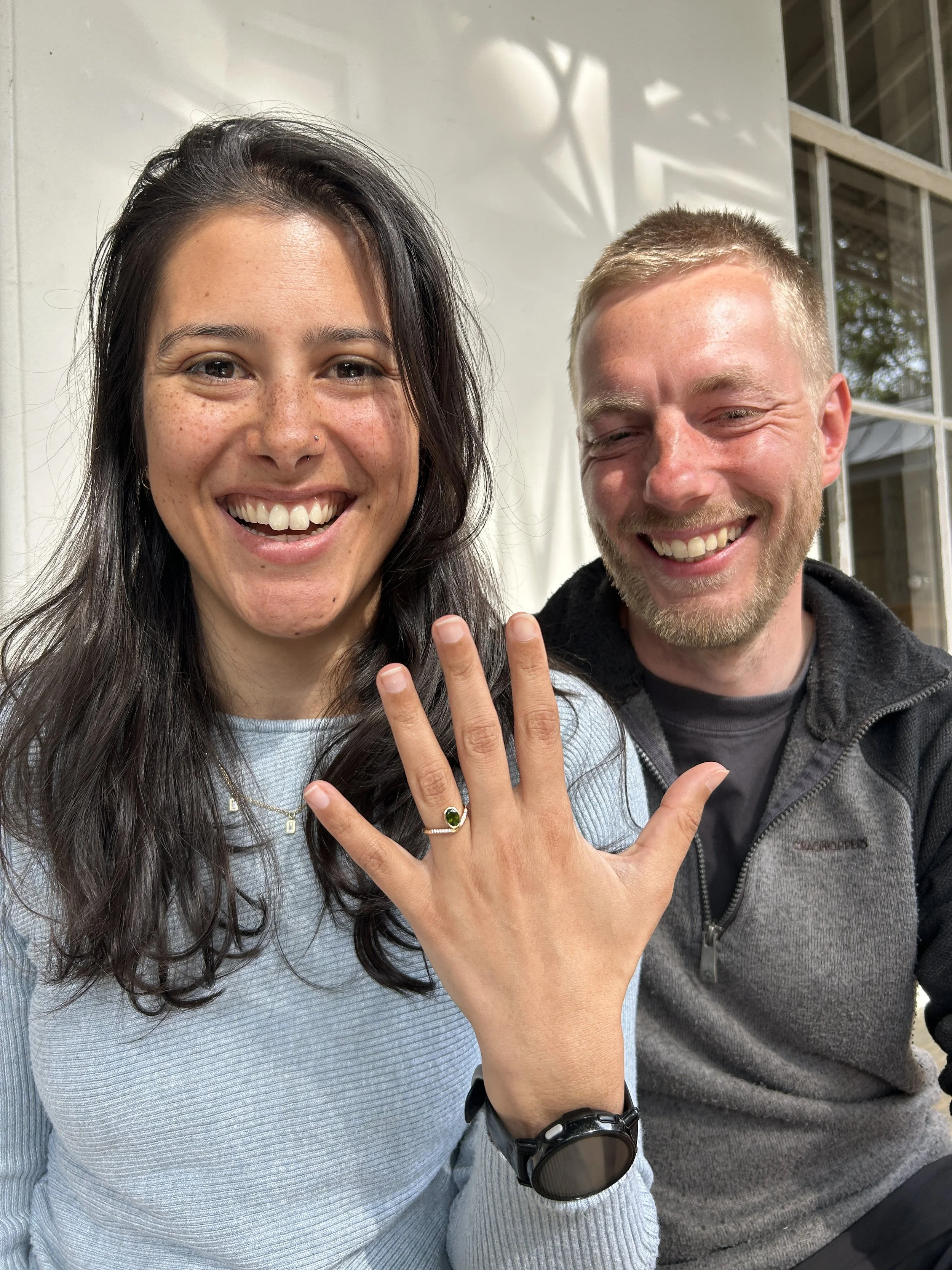 Close-up of a smiling woman with dark hair, freckles, and a nose piercing, showing her painted nails and a green ring, while a man with a beard and short blond hair smiles beside her, with their hands raised showing a peace sign.