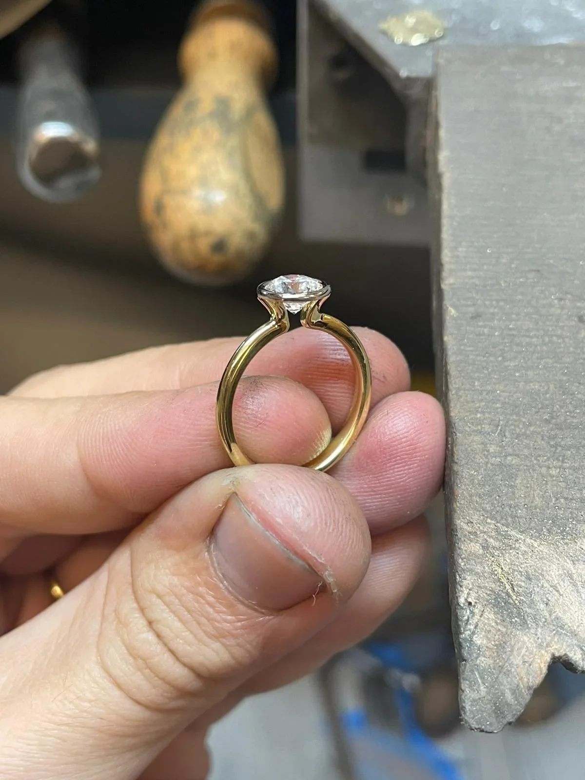 Close-up of a person holding a gold ring with a diamond in a jewelry workshop, with tools and a workbench in the background.