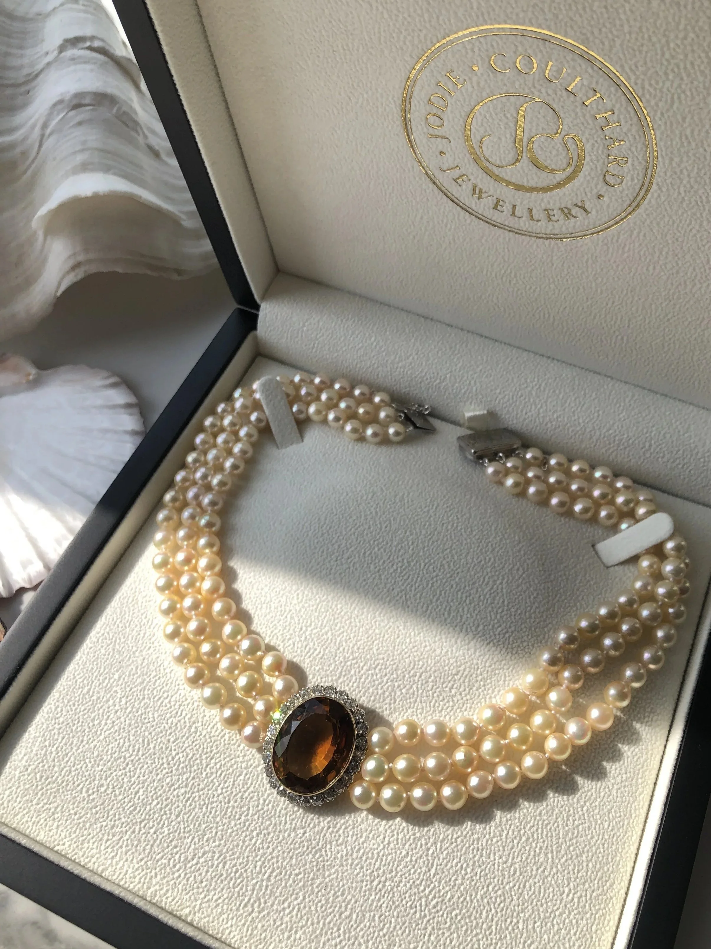 A jewelry box containing a pearl necklace with a large amber-colored gemstone pendant, with a jewelry logo on the lid that reads 'Jodie Marie Couture Jewelry'.