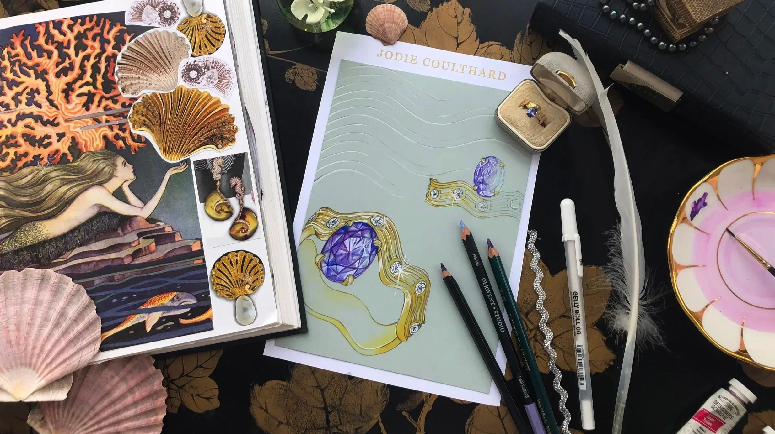 A jewelry display with various gold and gemstone jewelry designs, art supplies including colored pencils, printed illustrations of jewelry, and decorative shells on a black and gold surface.