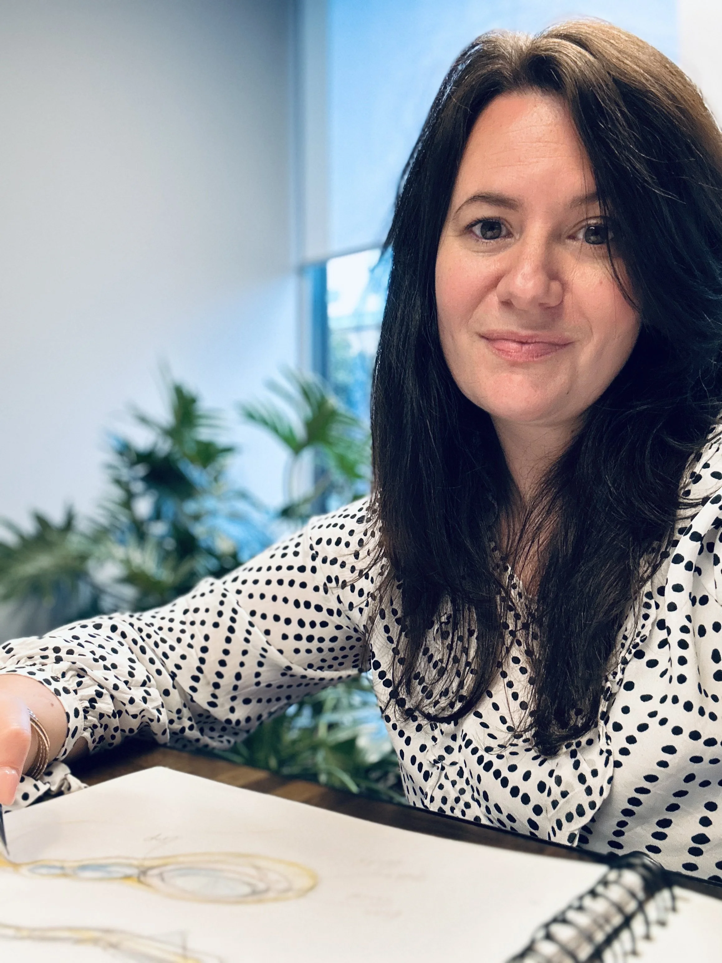 A woman with dark hair and wearing a polka dot blouse sitting at a desk with a notebook, colorful glasses, and a floral or leafy plant in the background.