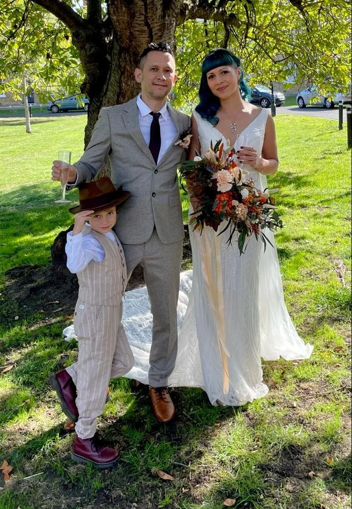 A wedding scene outdoors under a tree, featuring a bride and groom with a young boy. The groom is in a light gray suit holding a champagne glass. The bride is in a white wedding dress holding a bouquet of flowers. The boy, dressed in a 1920s-style striped suit and hat, is standing next to the groom, adjusting his hat with his hand.