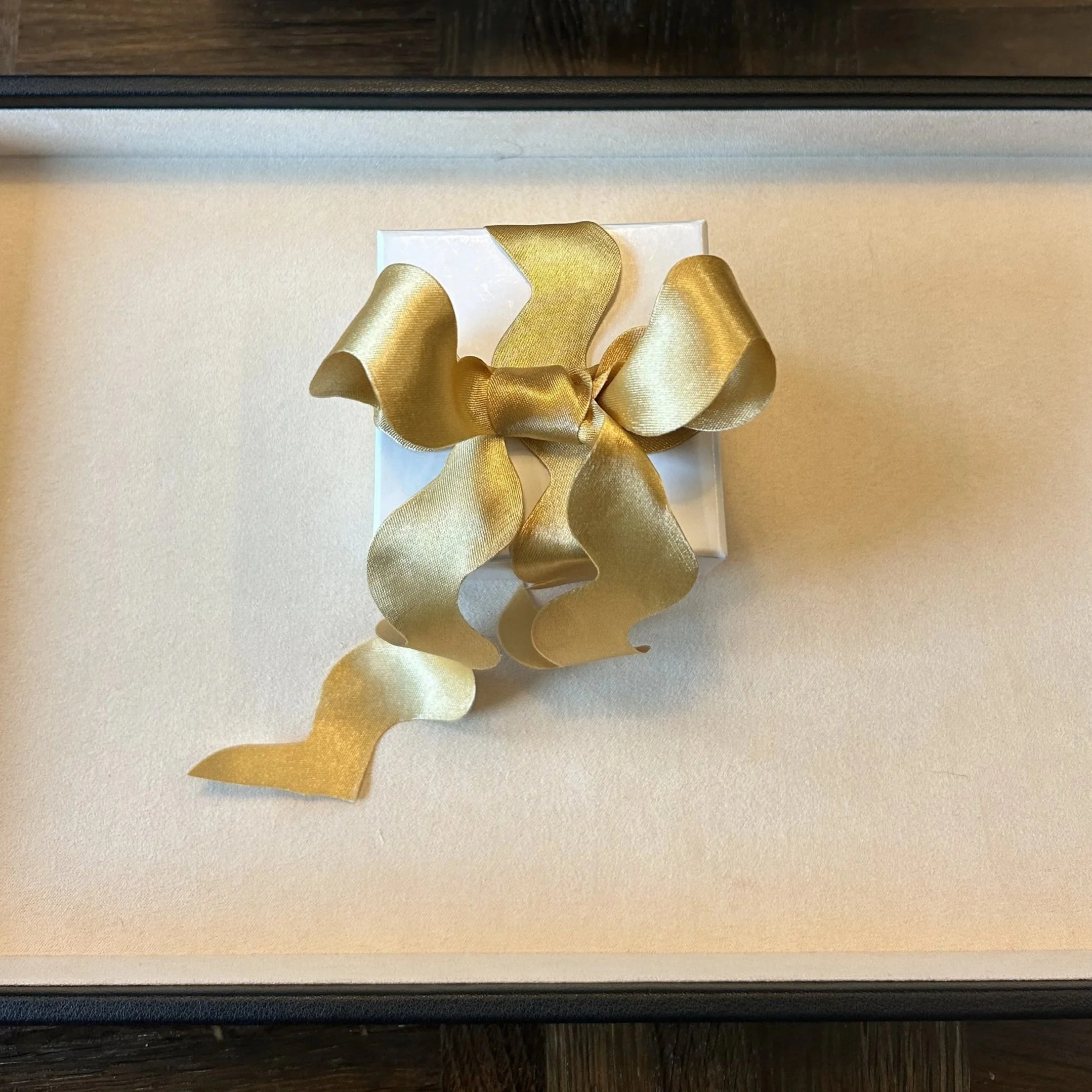 A Jodie Coulthard Jewellery wrapped gift ring box with a vintage satin ric rac gold ribbon bow on top, placed on a suede tray on a dark wooden table, ready for the customer to collect.