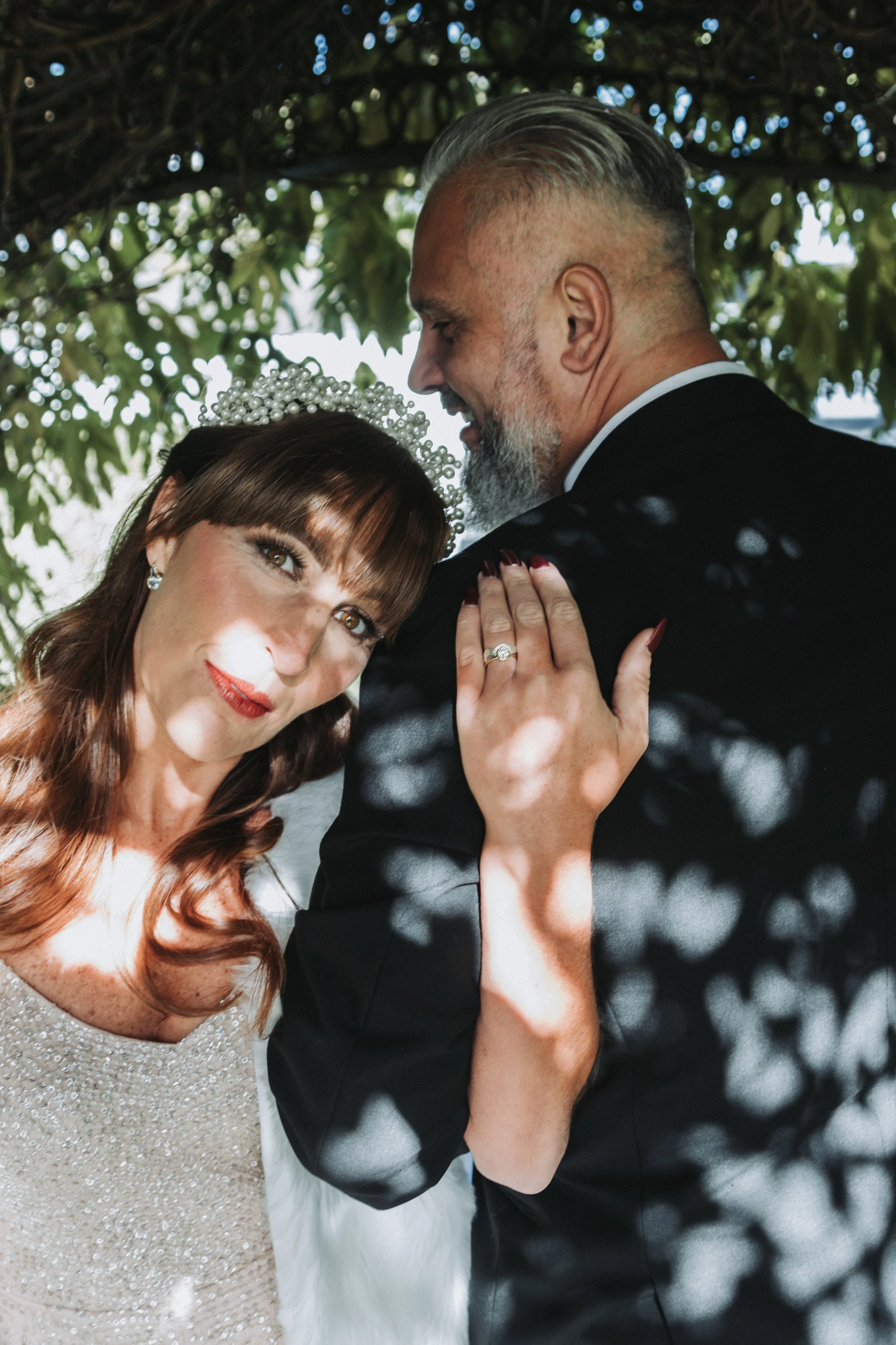 A couple dressed in formal attire, with the woman wearing a tiara and the man in a suit, sharing an intimate moment outdoors beneath a leafy canopy. Adorned with bespoke comfort wedding bands inspired by seashore & 1ct Natural diamond engagement ring