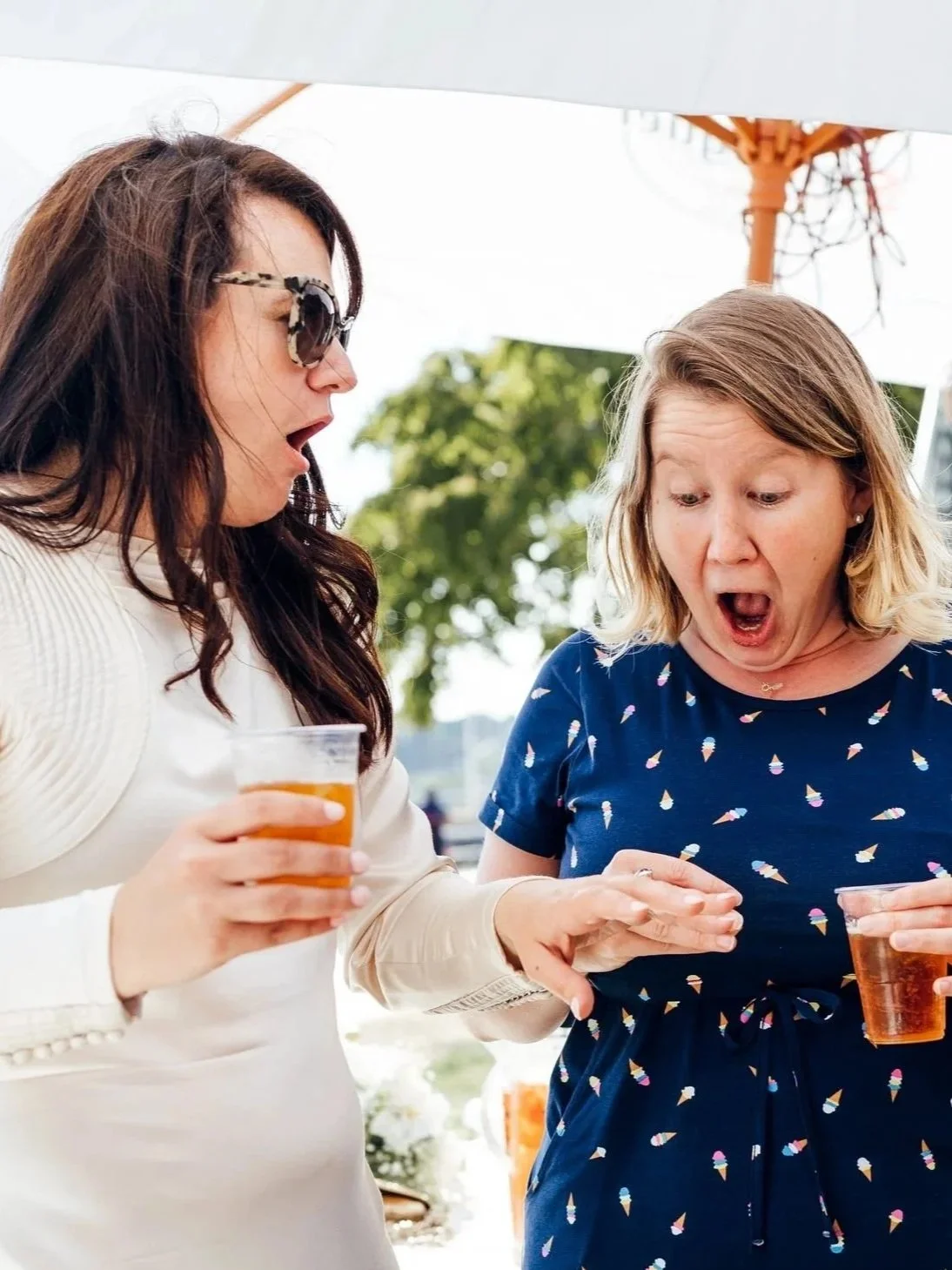 Two women at a wedding, holding drinks, with surprised expressions looking at a beautiful sapphire engagement ring.