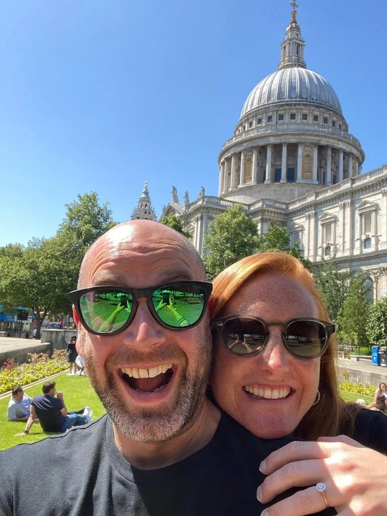 A smiling man and woman wearing sunglasses taking a selfie in front of St. Paul's Cathedral in London, with people relaxing on the grass nearby and clear blue sky overhead.