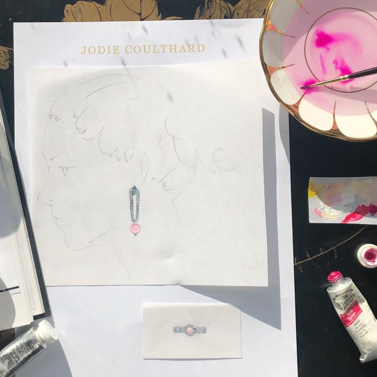 A sketch of a woman's face with a focus on earrings, signed by Jodie Coulthard, surrounded by painting supplies including pink paint and brushes, on a white table.