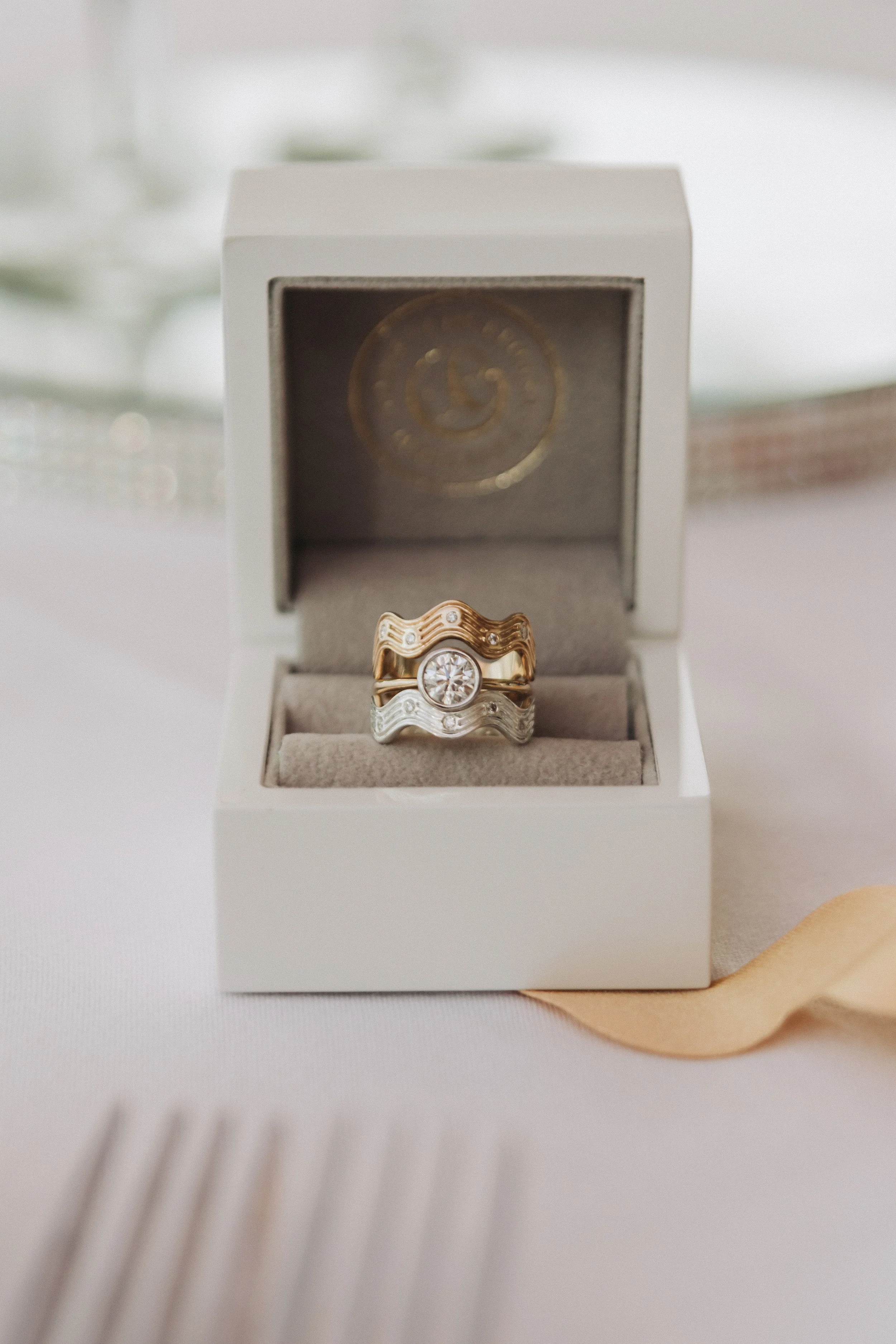 A white jewelry box with a ring inside, featuring a gold band with a large central diamond and smaller diamonds on the sides, placed on a white surface with a blurred background. Featuring waved wedding band flanking a 1ct diamond engagement ring.  