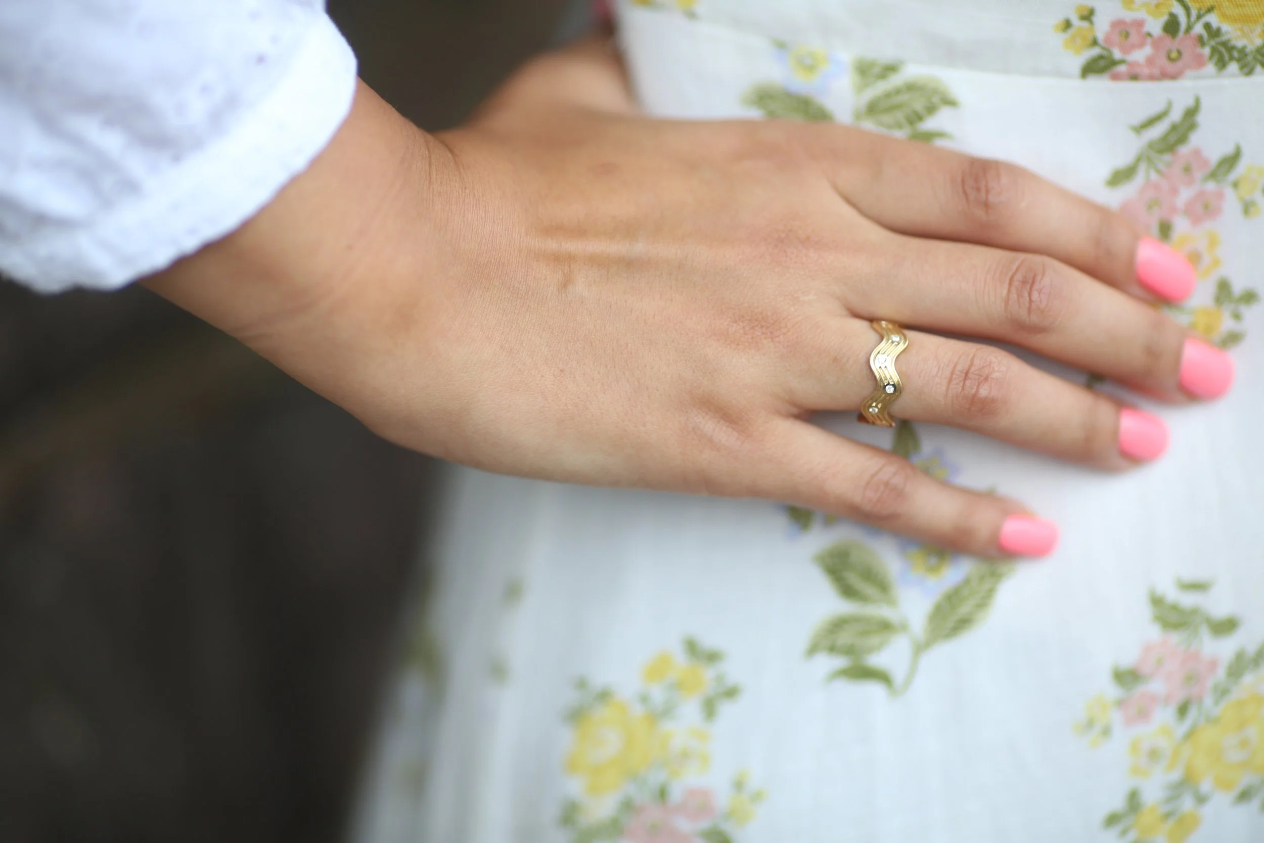 Close-up of a person's left hand resting on a floral-patterned fabric, showing a gold ring with a black and white design on the ring finger, and nails painted pink.