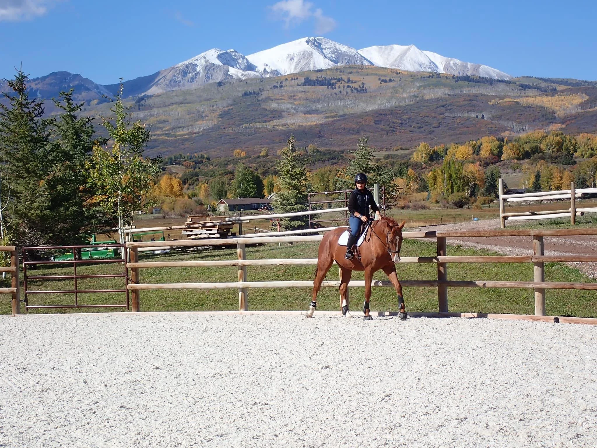 A woman riding a brown horse in an outdoor riding arena with a wooden fence, mountains with snow, trees, and a blue sky in the background.
