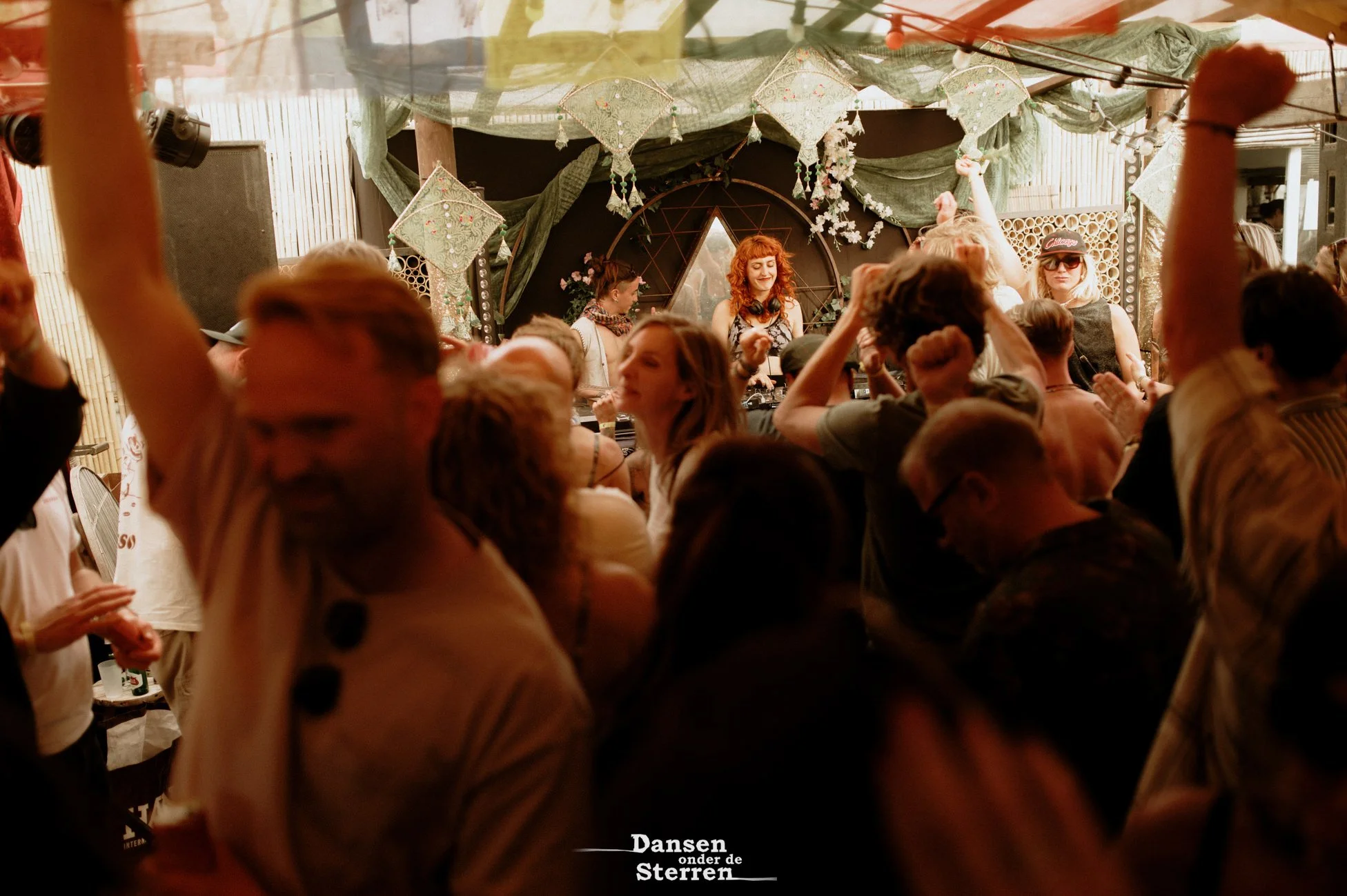 A lively dance party with a crowd of people dancing and socializing. In the background, a DJ is playing music, and decorated green fabric and paper flowers hang from the ceiling, creating a festive atmosphere.