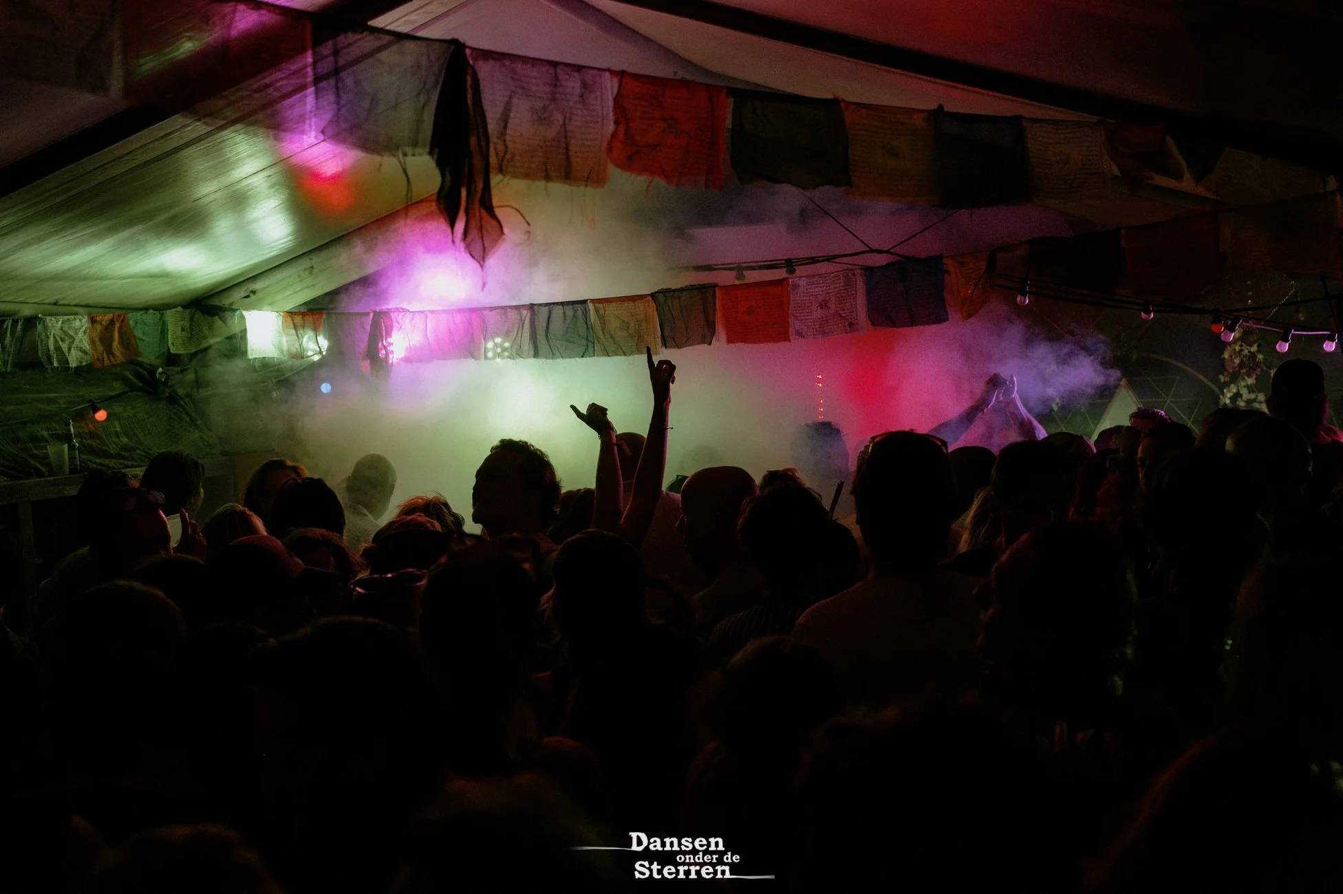 People dancing at a nighttime event with colorful lights and smoke effects, colorful cloth decorations hanging above, and a crowd watching the performance.