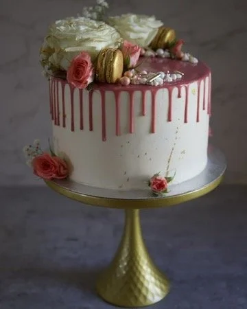 A white cake with pink drip icing, decorated with pink roses, white flowers, gold macarons, and silver sprinkles, on a gold cake stand.