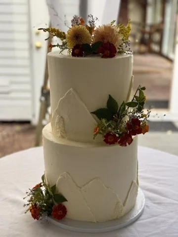 Two-tier wedding cake decorated with fresh flowers and greenery, set on a white table outdoors.