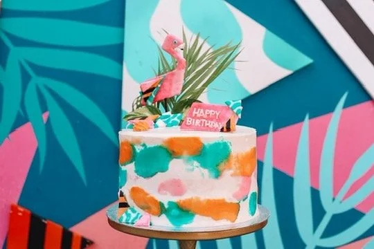 A colorful birthday cake decorated with pink flamingo and tropical-themed toppers, set against a vibrant tropical background with leaves and geometric shapes.