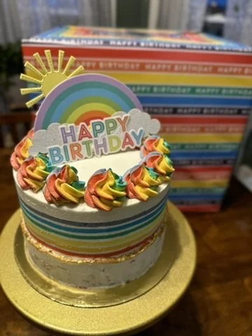 Rainbow-themed birthday cake with rainbow-colored frosting rosettes, a rainbow and sun topper, and a 'HAPPY BIRTHDAY' sign, placed on a gold cake board.