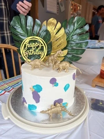 A birthday cake decorated with tropical green and yellow leaves, plastic dinosaur figures, and a yellow 'Happy Birthday' topper, on a table with a striped cloth.