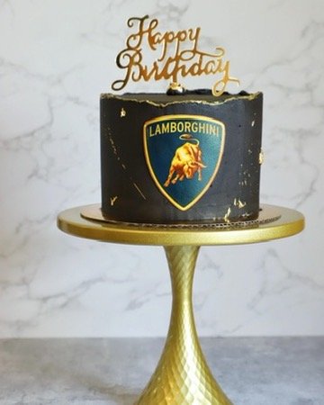 A black birthday cake with a Lamborghini logo, topped with a gold "Happy Birthday" cake topper, displayed on a gold cake stand against a light background.