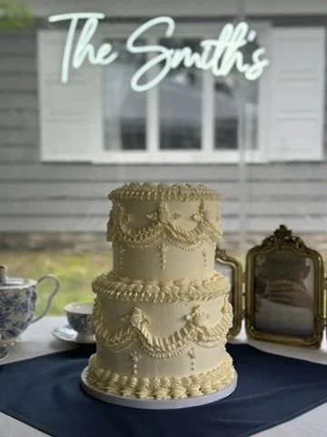 A three-tier wedding cake with white frosting and decorative piping, set on a table with a dark tablecloth, a blue and white teapot, and a mirror, with a window and sign that says 'The Smiths' in the background.