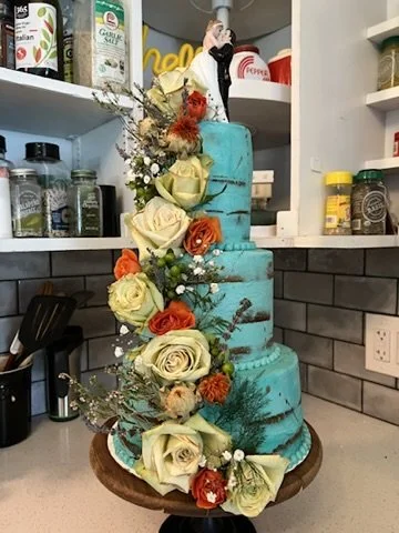 Four-tier teal wedding cake decorated with white, orange, and red roses and greenery, topped with a figurine of a bride and groom, placed on a kitchen counter.