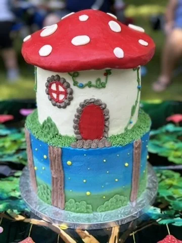 A whimsical cake designed to look like a fairy tale house with a red mushroom-shaped roof with white spots, a white walls, a red door, and a grassy base, set on a decorative cake board.